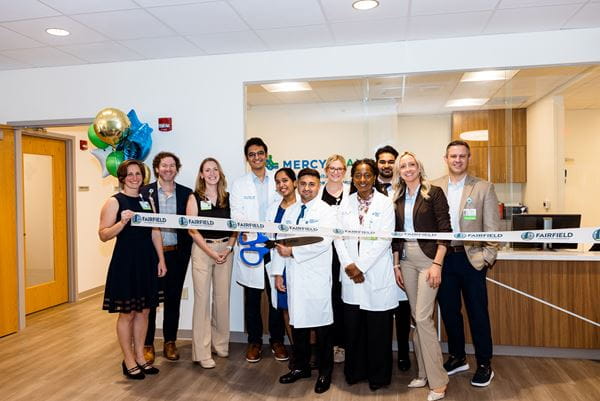 A group of eleven people, several wearing white medical coats and ID badges, stands together behind a "Fairfield Chamber of Commerce" ribbon during a ribbon-cutting ceremony, with one person holding oversized blue ceremonial scissors. Balloons and a Mercy Health sign are visible in the background of the modern medical facility.