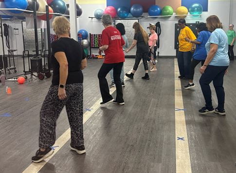 Dance fitness class at St. Joes At The Mall. Image of about a dozen people dressed in fitness clothes arranged in lines in dance studio.