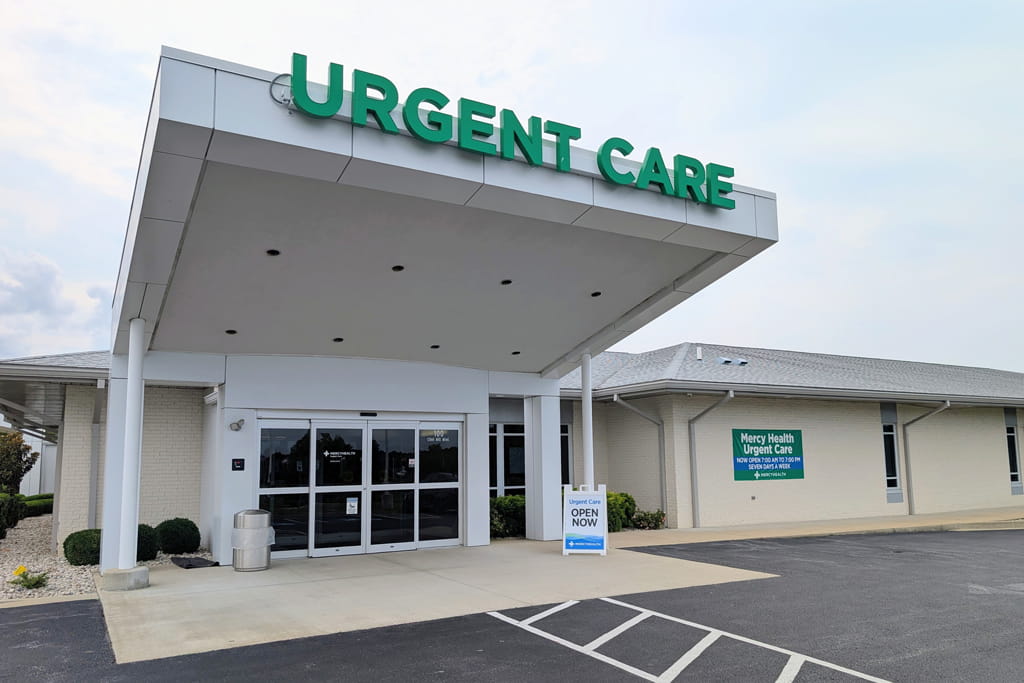 Exterior of Mercy Health Urgent Care in Paducah, KY, showing the entrance under a large canopy with a green 'Urgent Care' sign, an 'Open Now' sidewalk sign, and the parking lot in the foreground